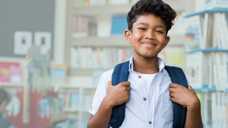 A smiling boy wearing a backpack stands in a library.