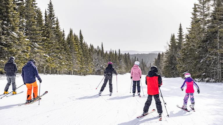 Kids and parents wearing colourful jackets ski on the snowy hill with trees around them.