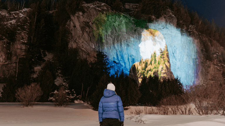 A woman in a dark blue winter jacket looks at the art being projected on a large rock during the evening.