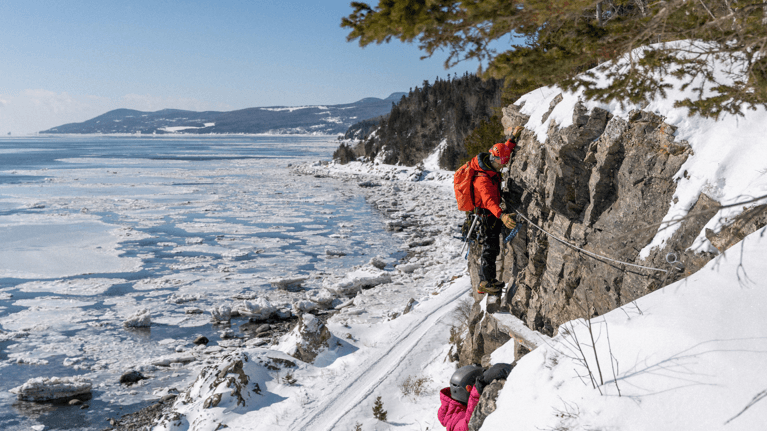 A father and his young daughter climb a large rock overlooking the frozen water and snowy mountains.