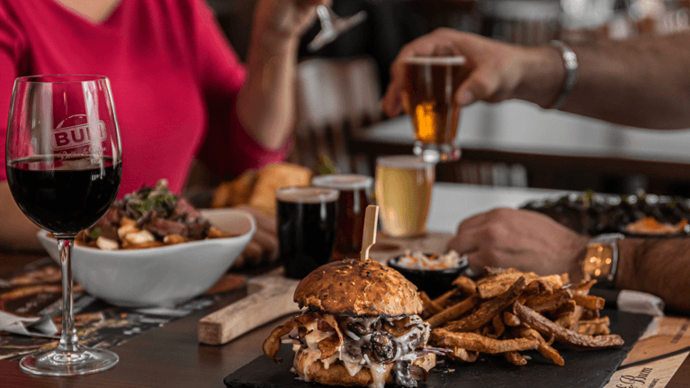 A large burger and fries sits on a table with a few other dishes and drinks.