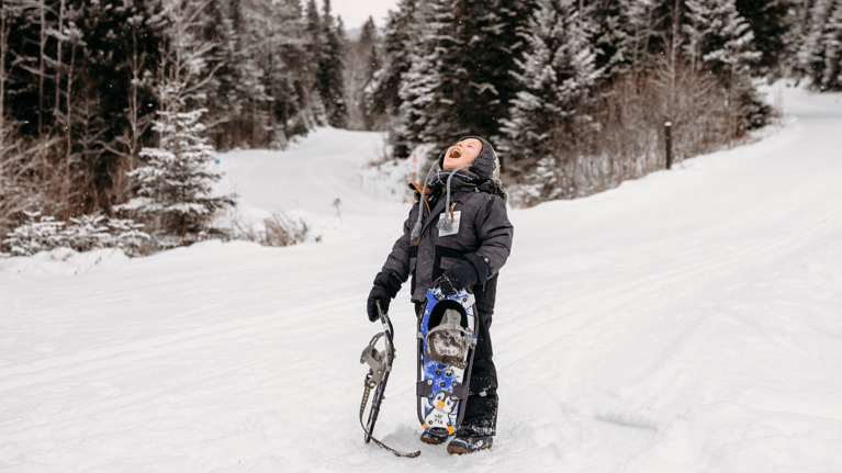 A young boy holds a pair of snowshoes and laughs as the snow falls down.