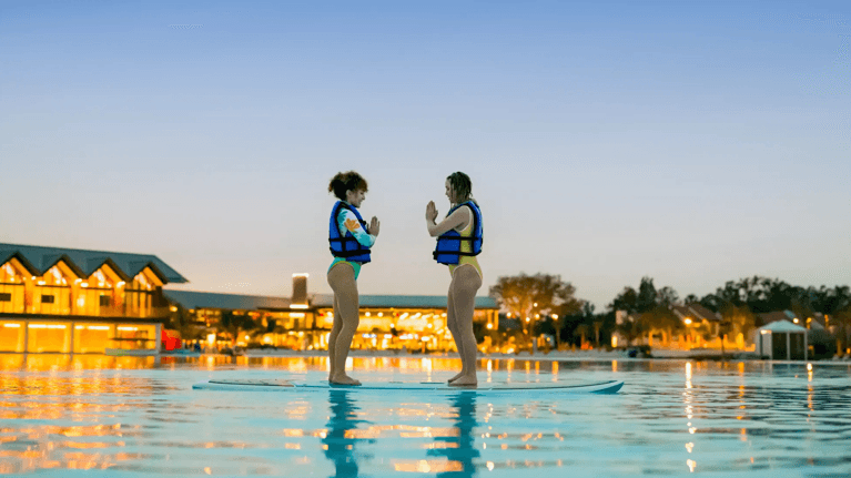 Two girls wearing swimsuits and life jackets stand on paddle boards, with warm lights from the buildings behind them reflecting on the water.