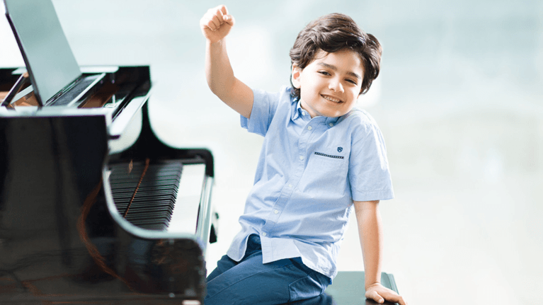 A little boy wearing a light blue shirt sits by the piano and cheers with his fist in the air.