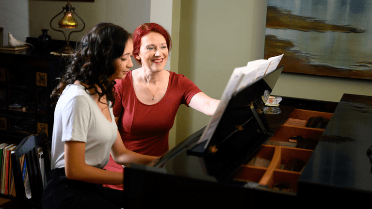 A piano teacher with a red t-shirt and red hair teaches a teenage girl how to play piano.