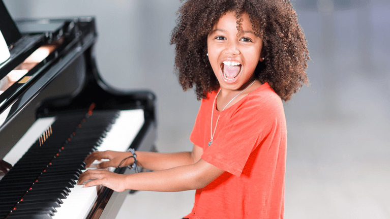 A young girl with curly hair plays the piano and smiles with her mouth open.