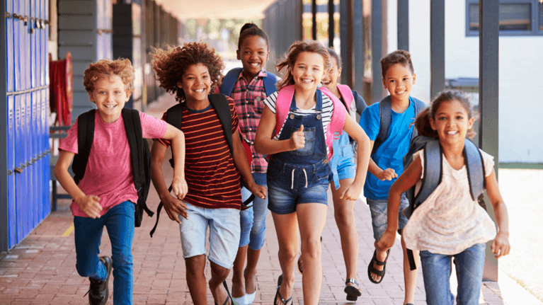 Kids wearing backpacks run in an outdoor school hallway.