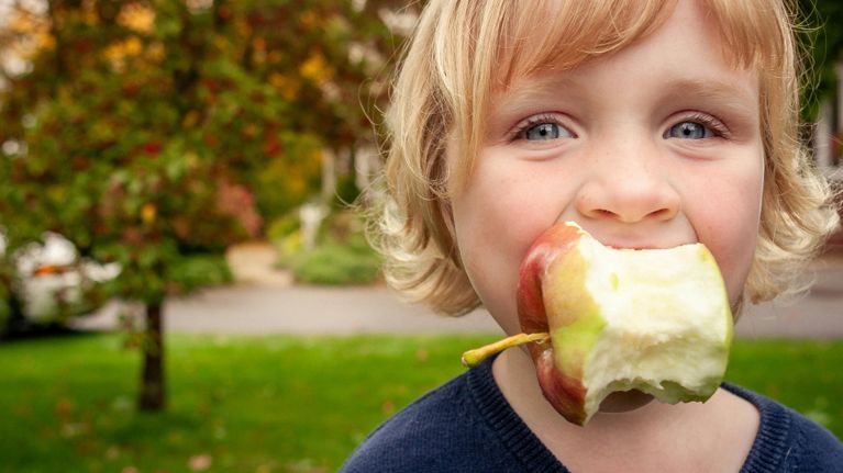 Close-up of a young child outdoors taking a big bite of an apple, with green grass and autumn-coloured trees blurred in the background.