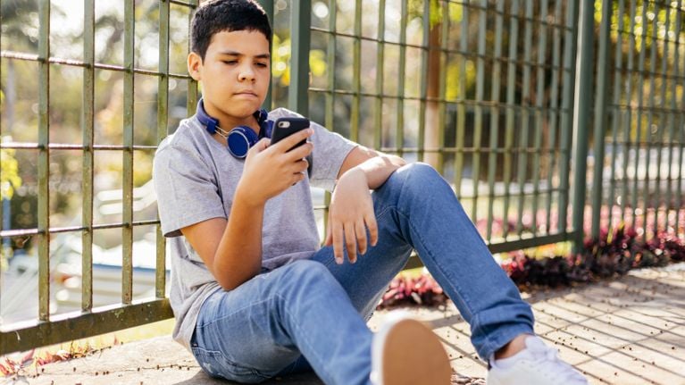 A tween boy wears headphones around his neck. He looks at a smartphone in his hand.