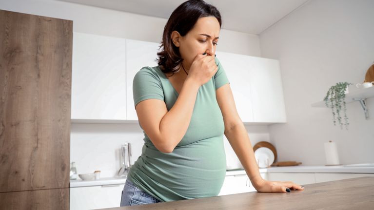 A pregnant woman standing in a kitchen, covering her mouth with her hand as she feels nauseous.