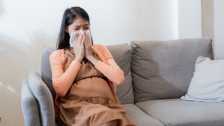A pregnant woman sitting on a couch sneezing into a tissue, wearing a peach cardigan and brown dress.