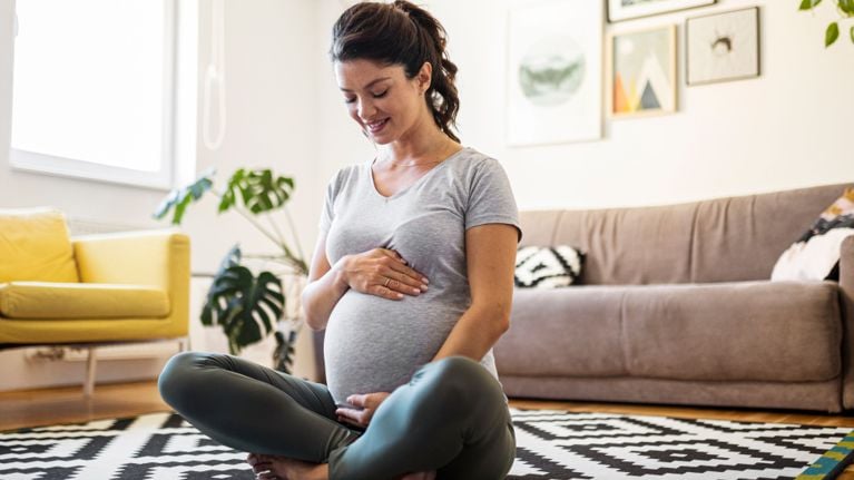 A pregnant woman sits cross-legged on the floor in a living room, gently resting her hands on her belly and smiling down at it.