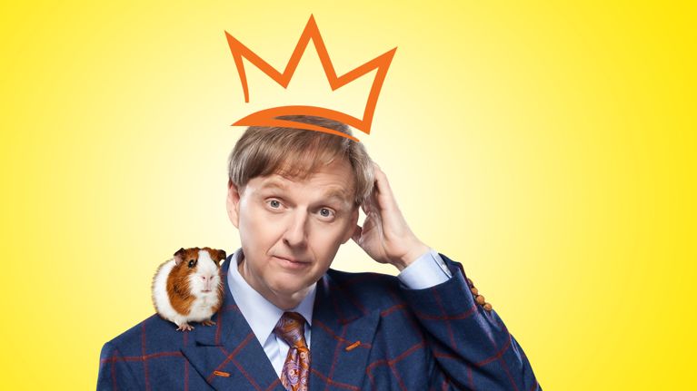 A photo of comedian Mac King wearing a suit with a drawn orange crown on his head in front of a yellow backdrop