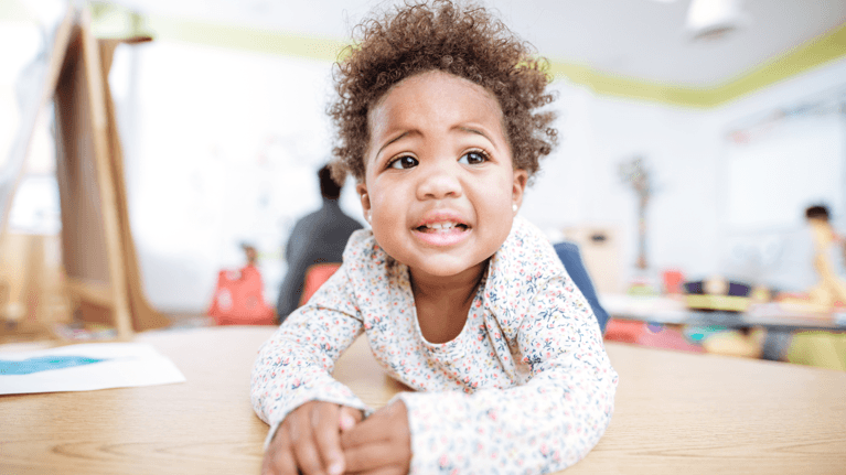 A toddler with curly hair leans on a table in a bright classroom, looking unsure or slightly worried.