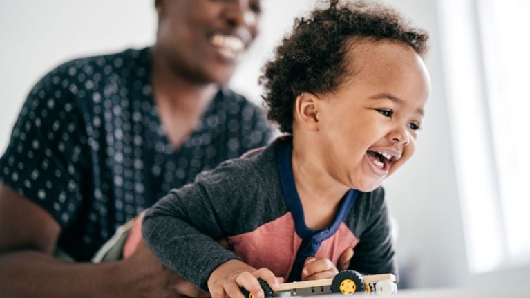 A laughing toddler lies on their stomach holding a small toy car while an adult behind them smiles and plays along.