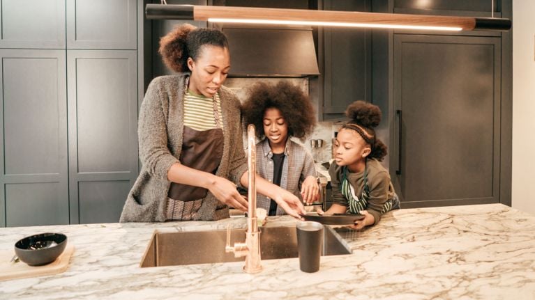A parent and two children stand at a kitchen island, washing dishes together at the sink in a modern kitchen