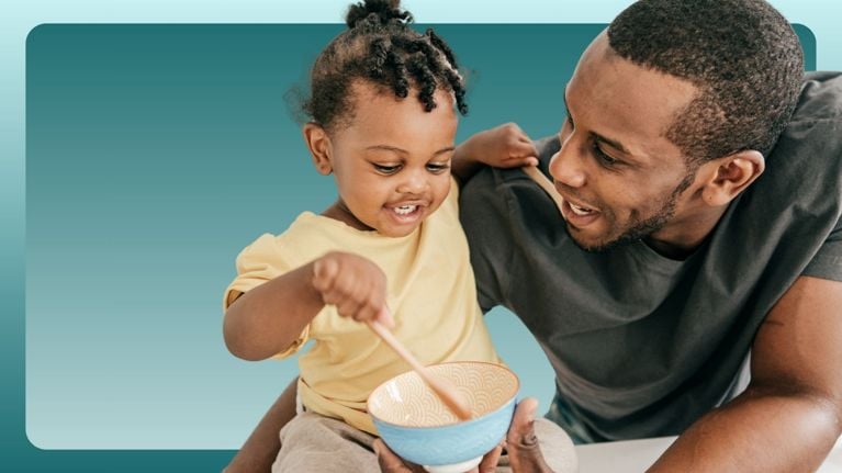 A smiling toddler stirs a bowl with a wooden spoon while sitting on a parent’s lap. Their joyful bonding moment shows how even babies and toddlers benefit from trust-building routines that introduce anatomical terms and reinforce privacy in a natural, everyday context.