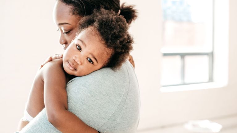 A caregiver holding a baby against their shoulder indoors, the baby resting their head calmly while looking toward the camera in soft natural light.