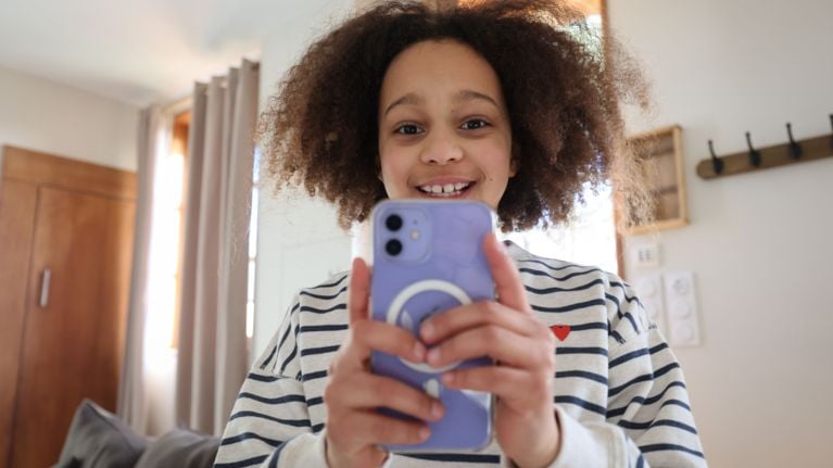 A young person is holding a smartphone in front of them and smiling at the camera. They are wearing a striped shirt with a small red heart emblem.