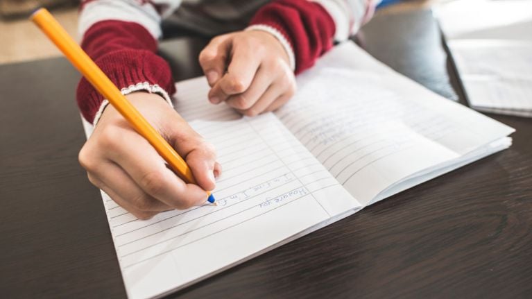 A person's hands are shown writing in a lined notebook with a yellow pencil. The writing appears to be practicing English phrases.