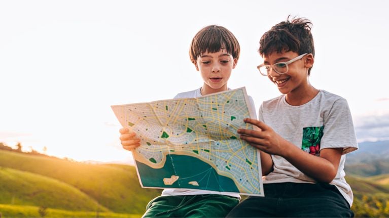 Two young people are sitting outdoors on a hillside and looking at a map together. One of them is wearing glasses and smiling.
