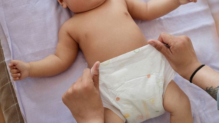 A caregiver changes a baby’s diaper on a soft, light-colored surface. The baby lies on their back with tiny fists curled, while the caregiver secures a fresh diaper with gentle hands.
