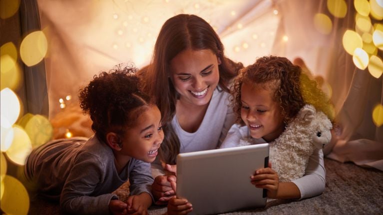 A woman with long, dark hair is seated in a makeshift tent made of blankets and fairy lights, looking at a tablet with two young girls. The girls, who have curly, dark hair, are smiling and engaged with the tablet's content. The cozy setting suggests a warm, intimate moment, possibly during a bedtime story or movie night. The overall tone is cheerful and loving.