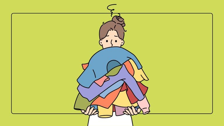 A person with a messy bun is almost completely hidden behind a towering stack of multi-colored folded clothes. Only their wide, slightly overwhelmed eyes are visible over the top of the laundry mountain.
