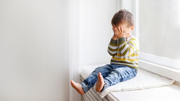 A young child sits on a window seat, their face hidden in their hands. The child is wearing a striped sweater and jeans and appears to be upset or overwhelmed.