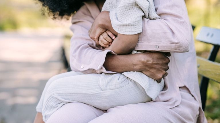 A close-up view of a woman warmly hugging a young child, both dressed in soft, textured clothing. They are sitting on a bench outdoors, with a soft-focus background suggesting a park or nature setting.