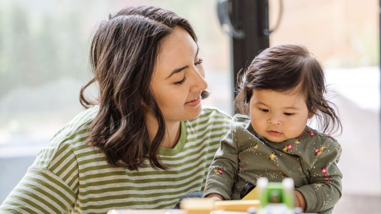A woman with long, wavy brown hair looks down at a young child sitting at a table. The child, with dark hair, is wearing a patterned long-sleeved shirt and appears to be focused on something in front of them.