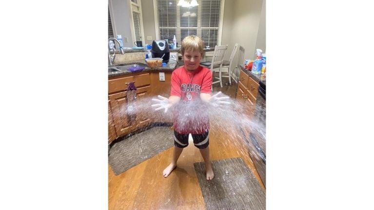 A young child with short blonde hair stands barefoot in a kitchen with their arms outstretched. Their hands are covered in a white powdery substance that is being thrown outwards in a burst. The child is wearing a red t-shirt with white lettering and black shorts with a white stripe. The kitchen features wooden cabinets, granite countertops, a stainless steel sink, and various household items in the background.