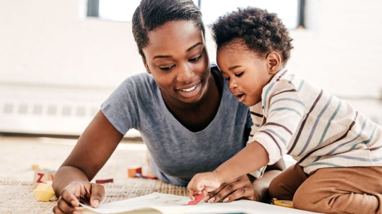 A woman and a young child are lying on a rug, looking at a book together. The woman appears to be pointing at something in the book, and the child seems engaged.