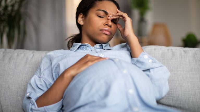 A pregnant person is sitting on a couch with their hand to their forehead. They seem to be experiencing discomfort.