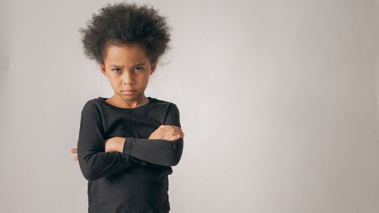 A young child standing with arms crossed and a defiant, angry expression, representing emotional resistance or frustration.