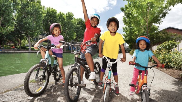 Four kids on bikes stand astride their bikes on a paved path.