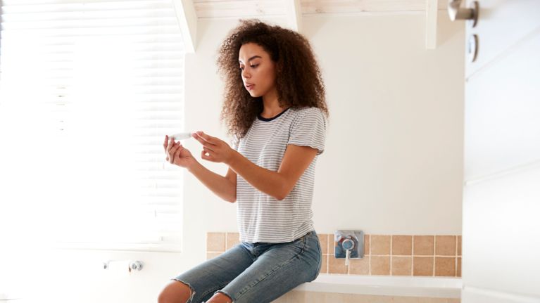 A young woman with curly hair sits on the edge of a bathtub in a softly lit bathroom, holding a pregnancy test and looking at it with a thoughtful expression. The image conveys the emotional weight and uncertainty that can accompany early signs of pregnancy.