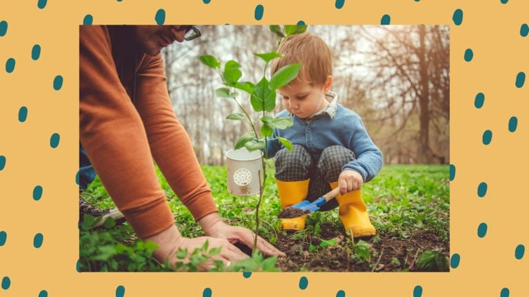 A young child in yellow rain boots and a blue sweater carefully scoops soil while planting a small tree with the guidance of an adult. They are outdoors in a grassy field, creating a memorial garden together under soft natural light.