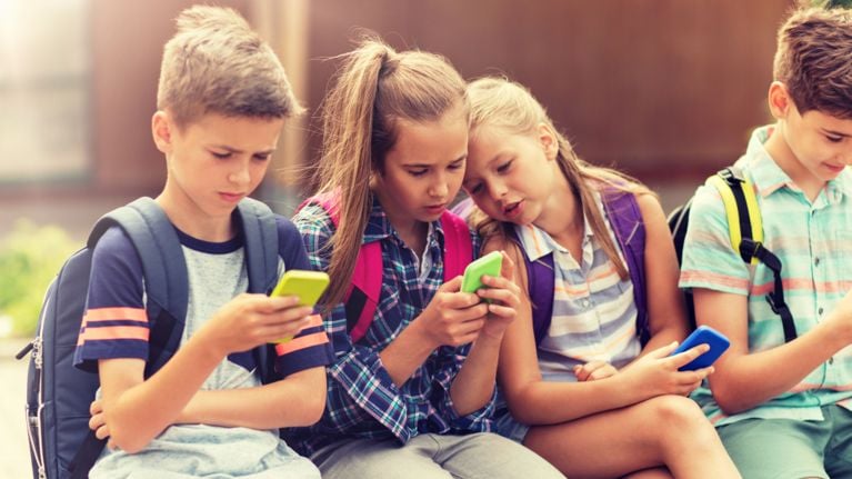 Several children are sitting together closely, each looking down at a smartphone. They appear to be focused on their devices.