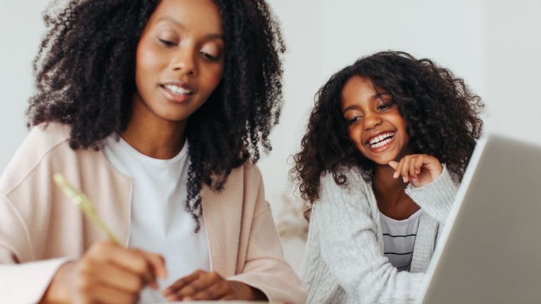 A mother writing in a notebook while her daughter smiles beside her at a laptop in a bright, minimal room.
