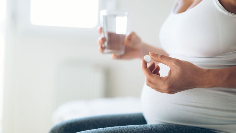 A pregnant person is holding a pill in one hand and a glass of water in the other. They are preparing to take medication.