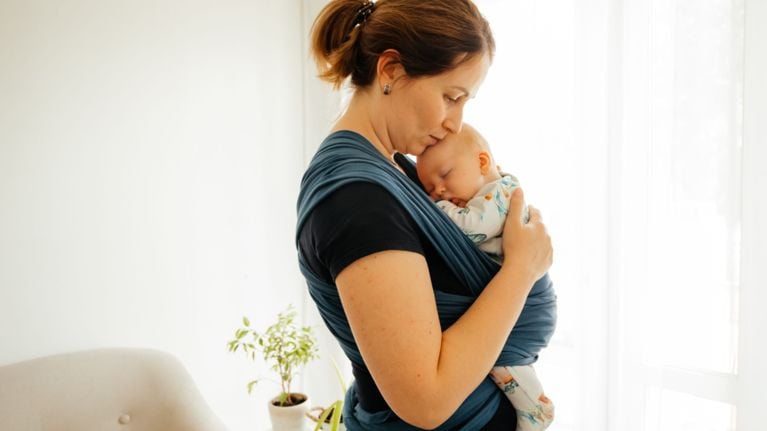A woman kissing the forehead of a sleeping infant wrapped in a blue baby carrier.