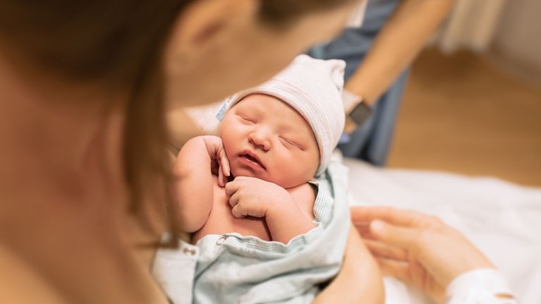 A newborn baby wrapped in a hospital blanket and hat rests peacefully in their parent’s arms shortly after birth.
