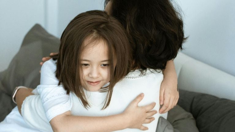 Two people, one older and one younger, are embracing each other tightly. The younger person is looking slightly downwards while being hugged.