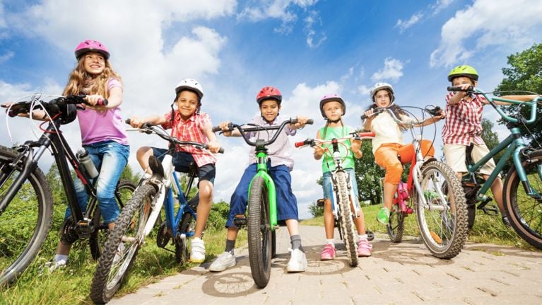 Several children wearing various colored helmets are riding bicycles together on an asphalt path. They are all smiling and looking forward.