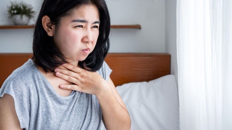 An adult woman sits on a bed, pressing a hand to her throat with a pained or uncomfortable expression. This image may represent nausea, gag reflex sensitivity, or increased saliva production—less common but possible early pregnancy symptoms.