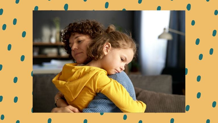A mother gently hugs her young daughter, who rests her head on her mother’s shoulder with a sad expression. They are seated indoors in a cozy living room, conveying emotional warmth and support during a moment of grief or sadness.