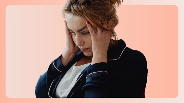 A tired mother in pajamas holds her head in her hands, eyes downcast in frustration and exhaustion. The image conveys the quiet intensity of mental overload and emotional fatigue many parents experience.