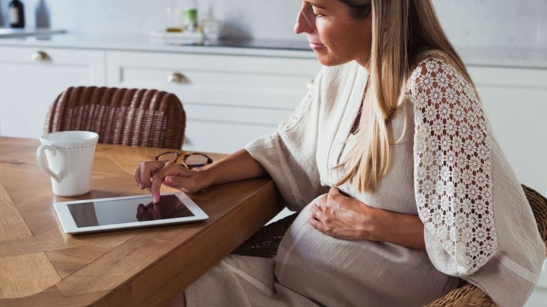 A pregnant person is sitting at a table with a tablet and a mug. They are looking at the tablet and holding their abdomen.