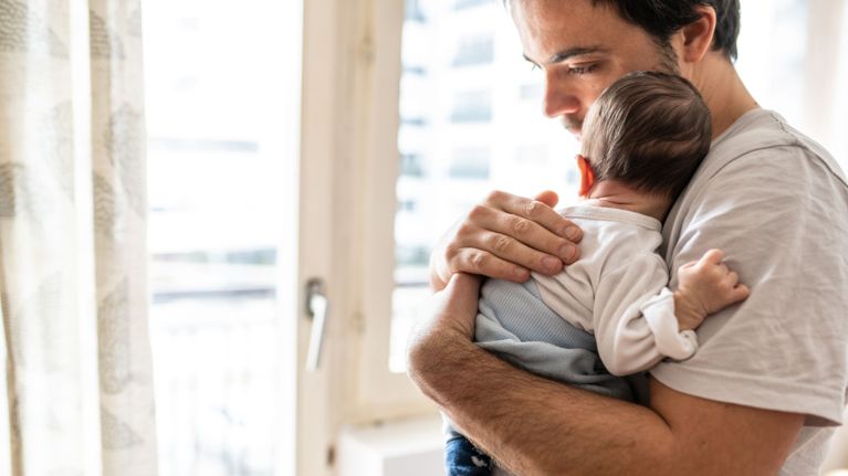 A father holding his newborn against his chest while looking out the window with concern.